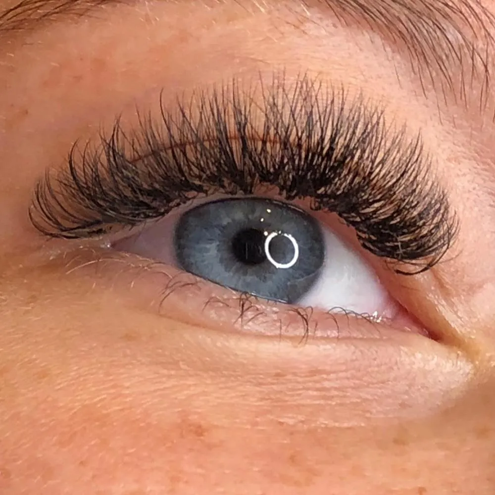 Wimpernverlängerung in Volumen: dichte, gefächerte Wimpern in weitem Schwung über einem blaugrauen Auge.